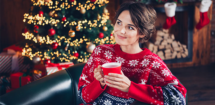 Frau im Weihnachtspulli sitzt mit einer Tasse in der Hand vor dem Weihnachtsbaum
